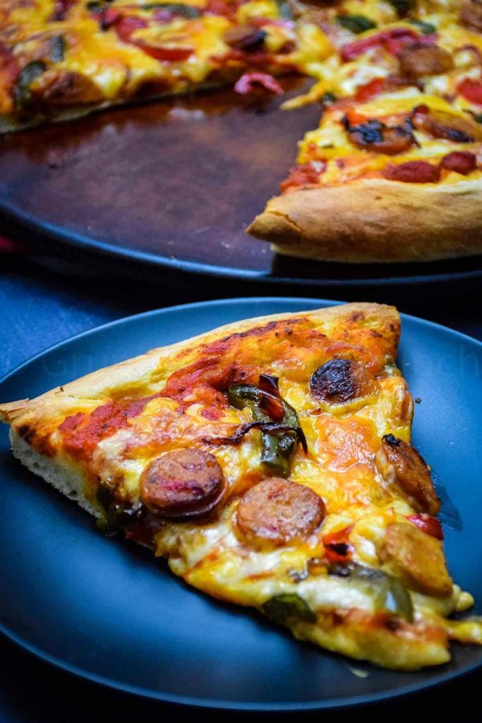 A slice of chicken sausage pizza on a black plate with the whole pizza in the background