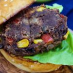 Close up of a spicy black bean burger patty with visible corn and red bell pepper pieces, served on a sesame seed bun with fresh green leaf lettuce.