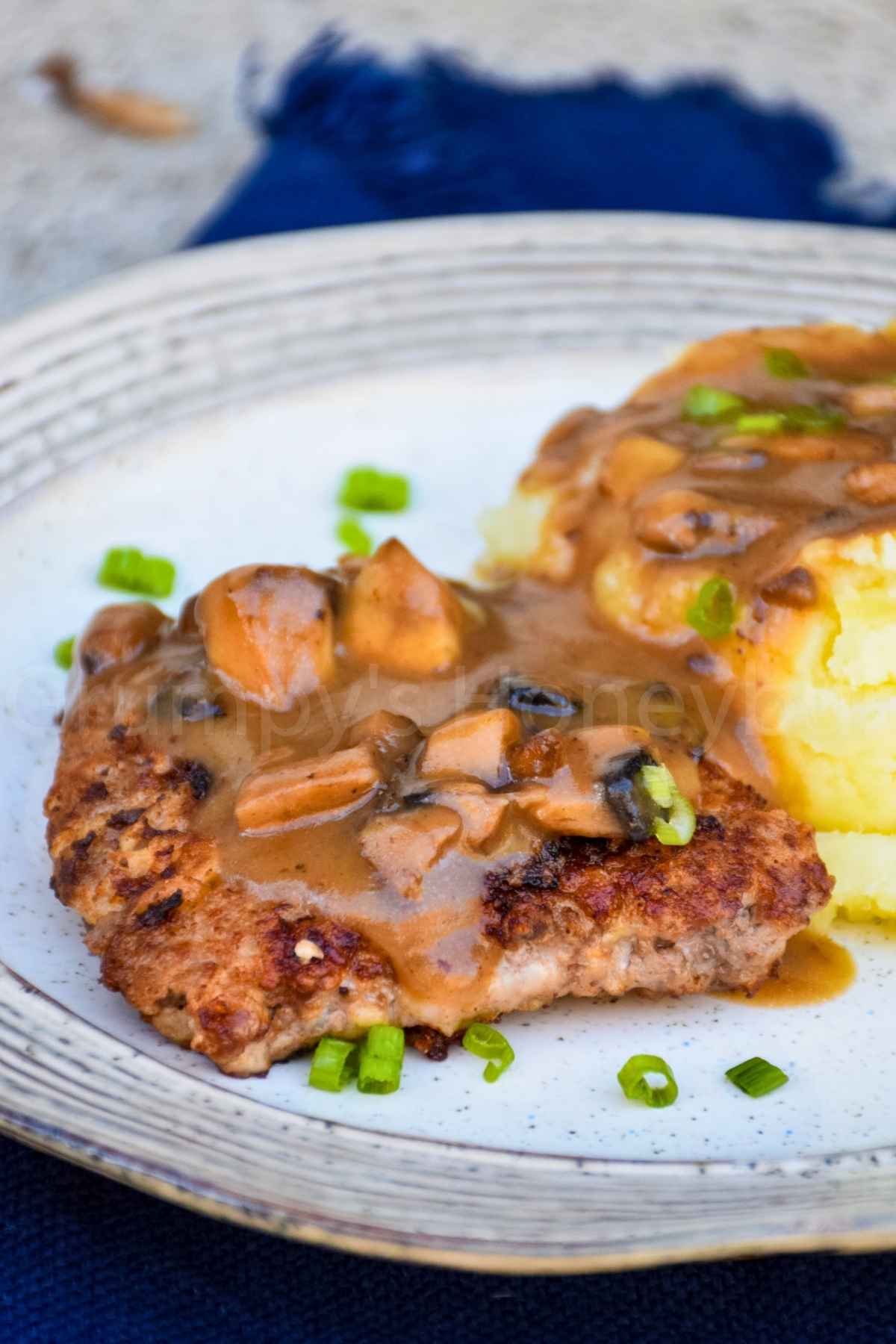 Close up of golden brown chicken fried steak, covered with a brown gravy studded with chopped mushrooms and a chopped chive garnish.