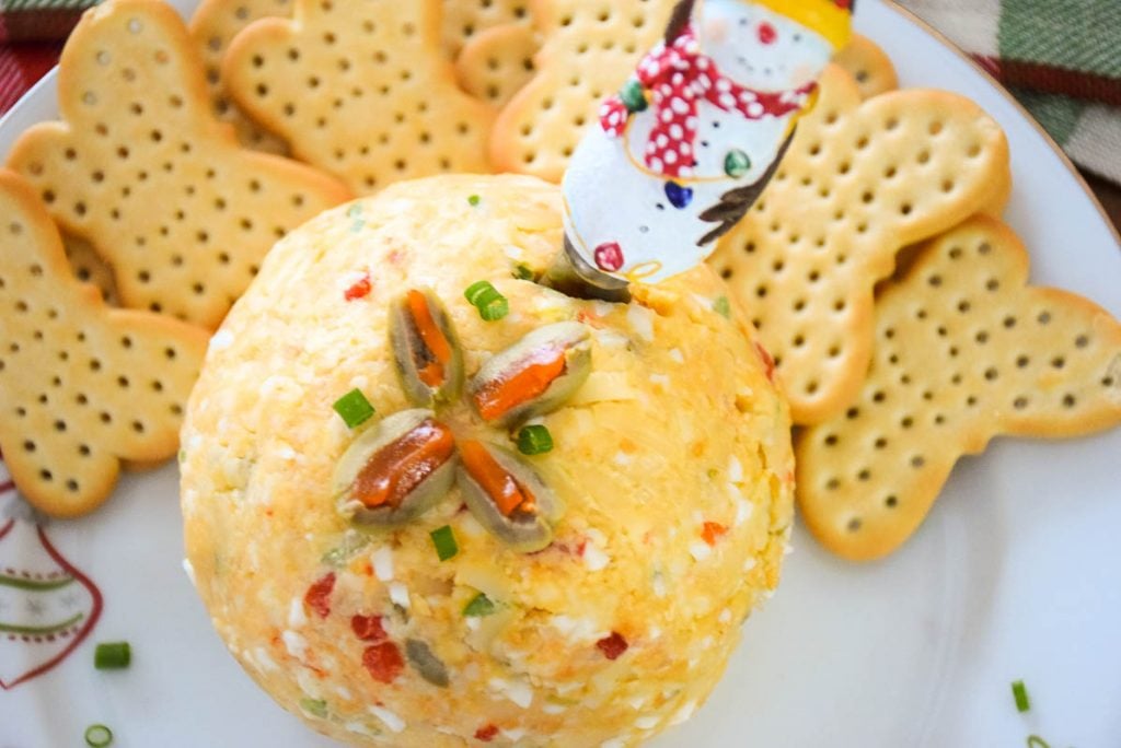 Overhead photo of easy cheese ball recipe with quartered green olives on top in a star shape and snowman cheese spreader in the cheese ball.