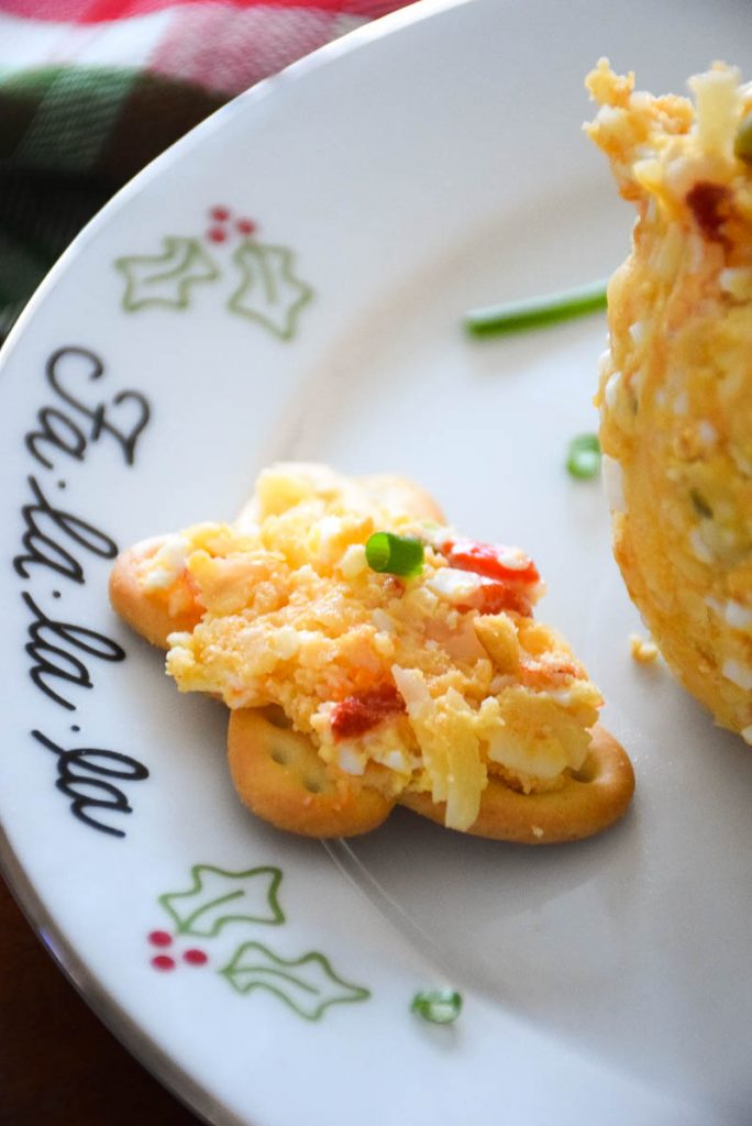 Mom's Easy Cheese Ball recipe spread on a buttery cracker on a white plate with holly print on the plate.