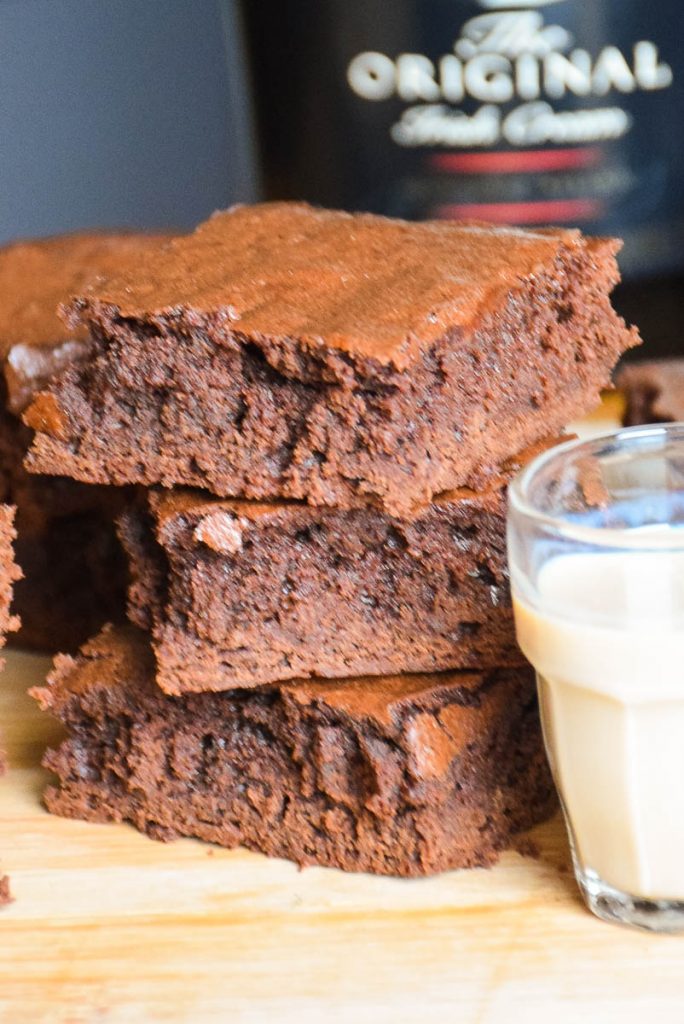 A stack of 3 baileys brownies with a shot glass of irish cream to the right and more brownies in the background.