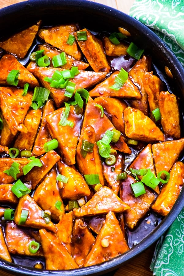 Overhead skillet view of Triangular shapes of crispy general tso's tofu coated in sauce and sprinkled with green onion tops.