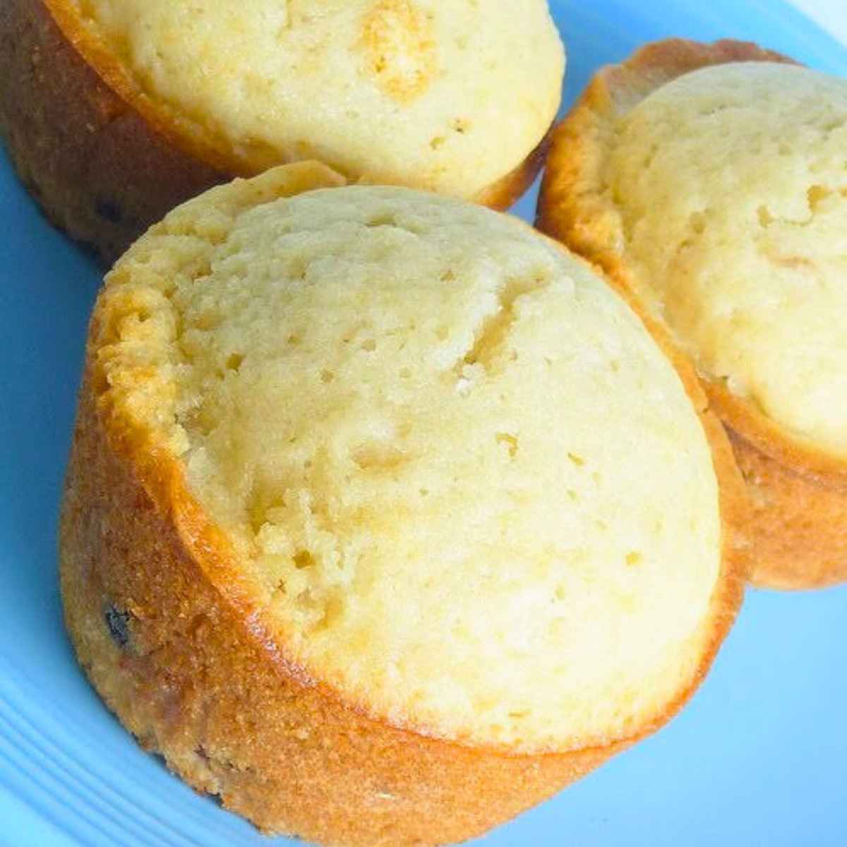 Three whole golden-brown rum raisin muffins with rounded tops arranged on a bright blue plate.