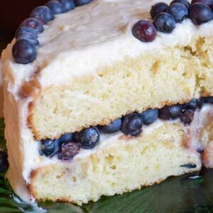 A close up slice of Banana Layer Cake showing a soft yellow crumb, a layer of fresh blueberries, and thick white lemon cream cheese frosting.