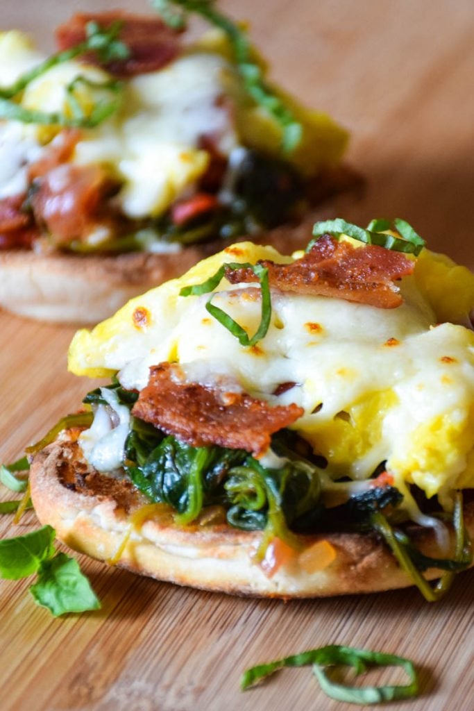 Breakfast Bruschetta in foreground made with a toasted english muffin topped with a tomato spinach base then layered with scrambled eggs, bacon, and cheese