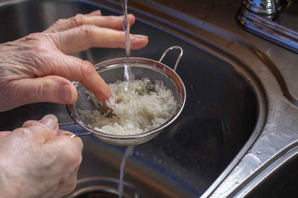 raw rice in a strainer being rinsed with water under the faucet