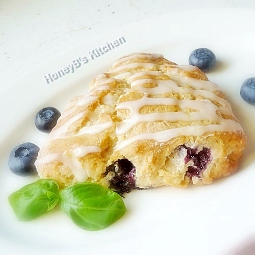 One blueberry scone on a white plate wit fresh blueberries scattered around it