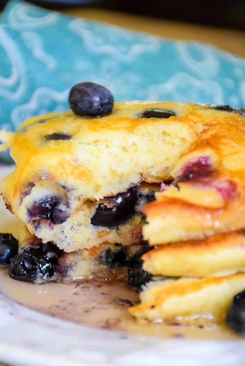 Three stacked pancakes filled with blueberries topped with maple syrup and a section cut out of the pancakes with a pool of syrup on the bottom of the plate and a blue napkin blurred in the background