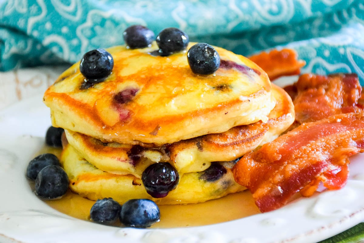 White plate with scalloped edges with a stack of 3 blueberry pancakes with a pool of maple syrup on the bottom of the plate, blueberries scattered on top of and around the pancakes and crispy cooked bacon to the right of the pancakes.