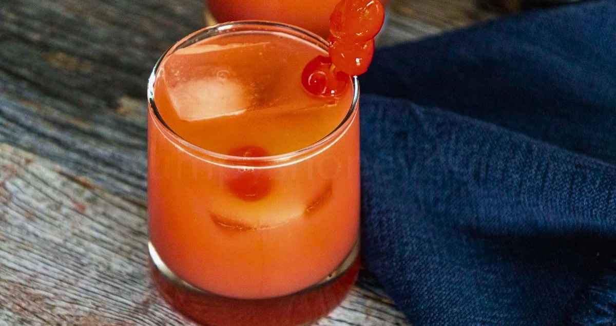 Upclose image of a reddish pink cocktail with ice and maraschino cherry garnish.