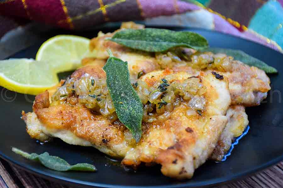 Sauteed Chicken Thighs topped with sage browned butter sauce, crispy sage leaves on a black plate with a sliced lemon on the side