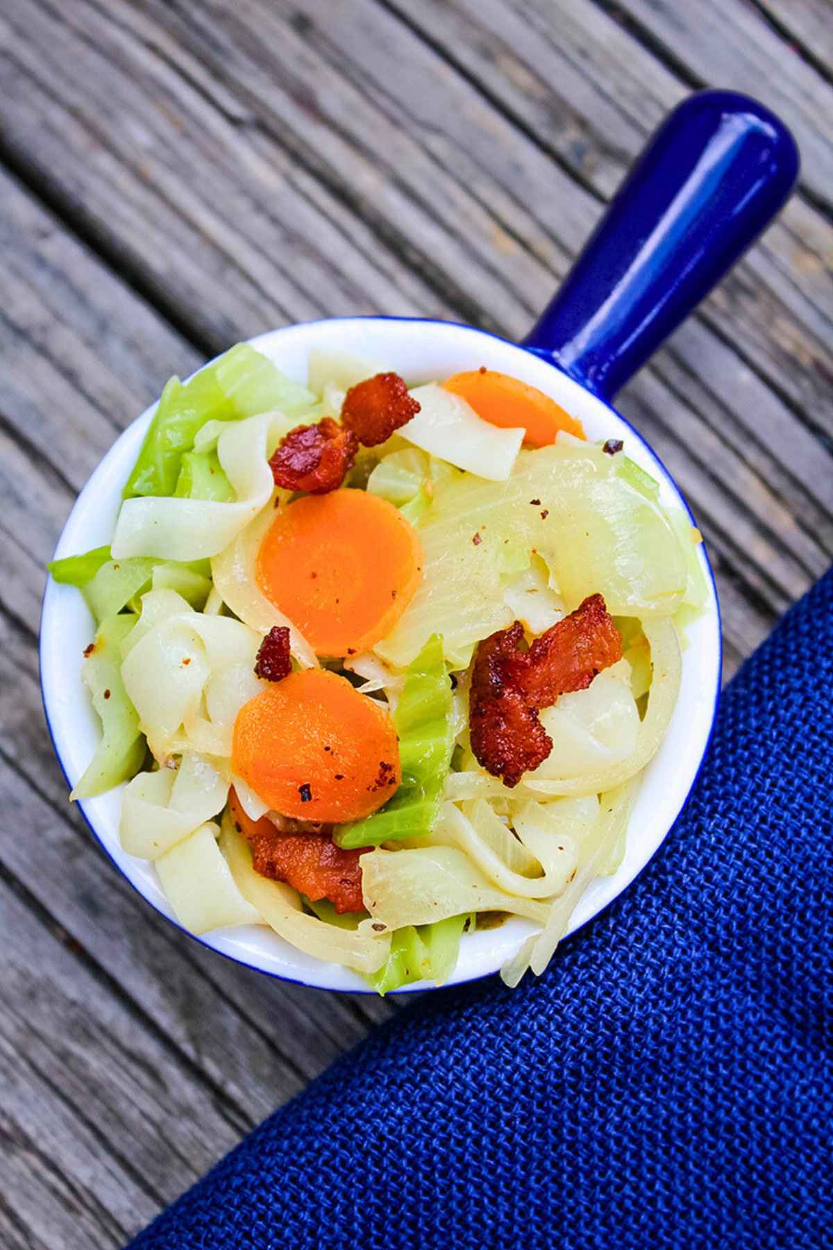 Overhead view of cabbage noodles with bacon, carrots, and onions in a white bowl with a blue handle.