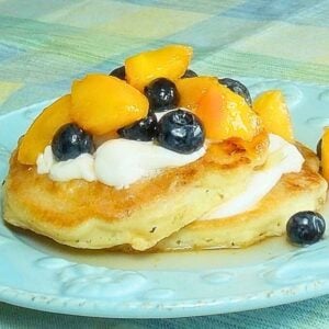 Close-up of fluffy pancakes topped with yogurt, peaches, and blueberries served on a light blue plate.