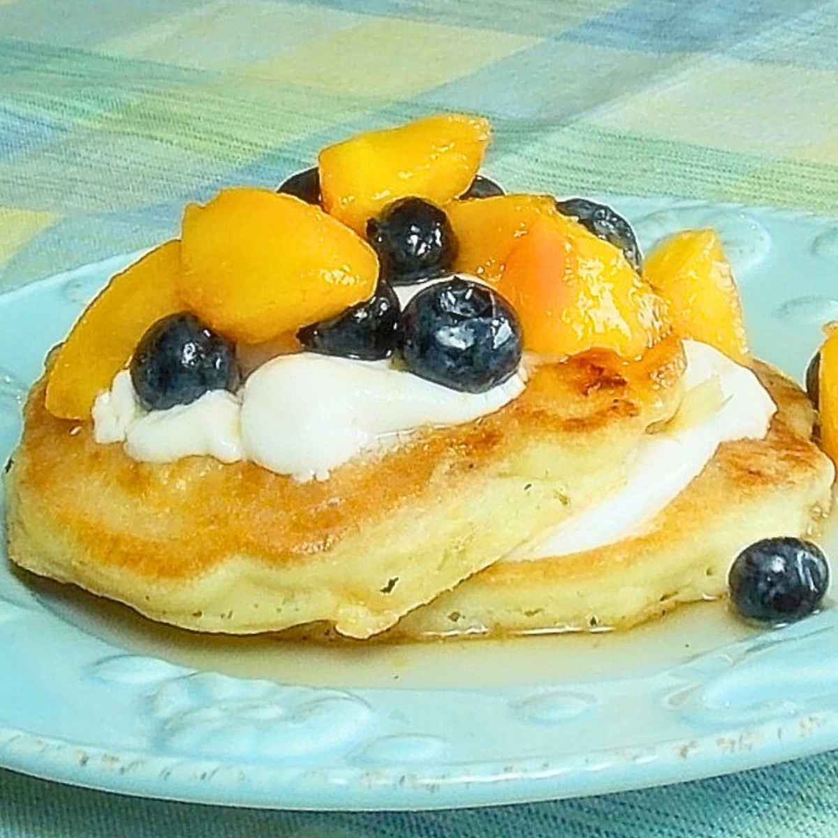 Close-up of fluffy pancakes topped with yogurt, peaches, and blueberries served on a light blue plate.