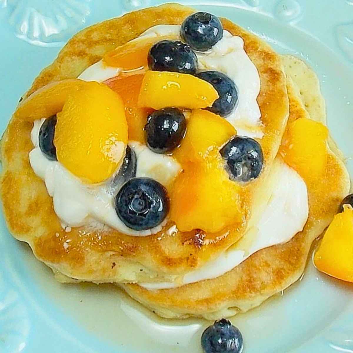 Overhead image of fluffy peach pancakes topped with yogurt, peaches, and blueberries served on a light blue plate.