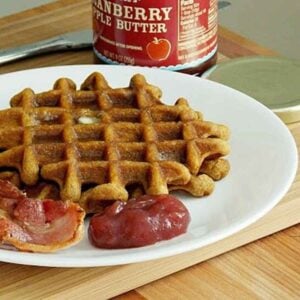 Two gingerbread wafflies on a white plate with a side of bacon and cranberry apple butter.