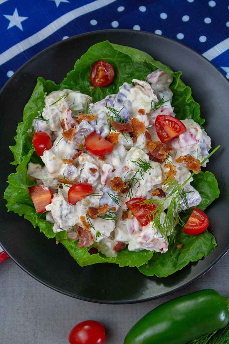 Overhead view of a serving of red white and blue potato salad on a black plate with lettuce beneath the salad. Jalapeno and grape tomato sitting at bottom of plate.