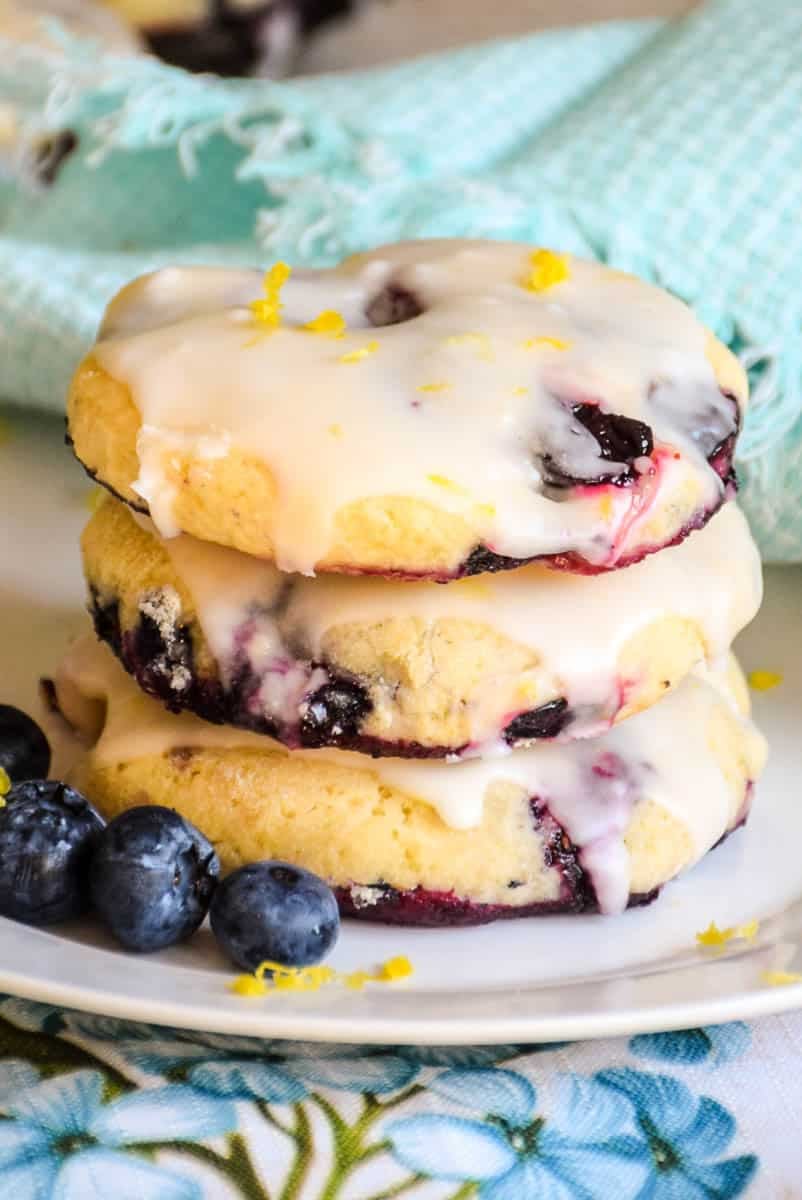 A stack of 3 lemon glazed cookies on a plate with a few fresh blueberries beside them and some lemon zest sprinkled over top.