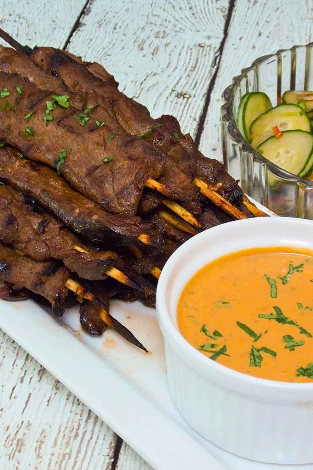Up close image of Venison Satay on a white serving tray with a side of spicy peanut sauce for dipping and a bowl of asian pickles.