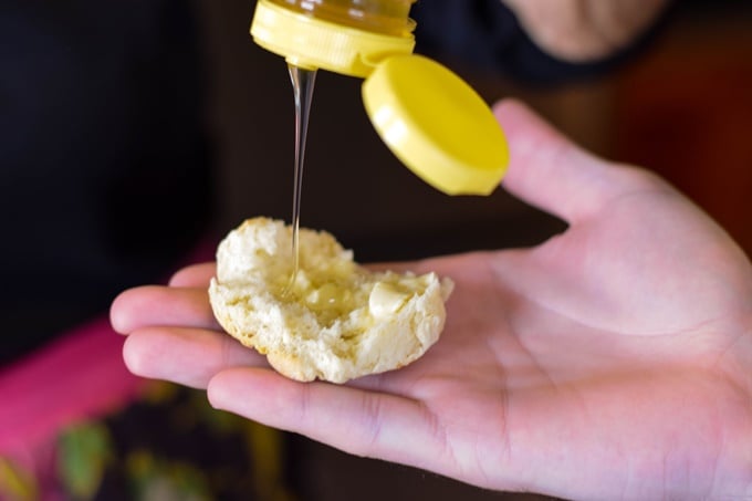 A hand holding an old fashioned buttermilk biscuit that was broken in half and honey drizzling over top.