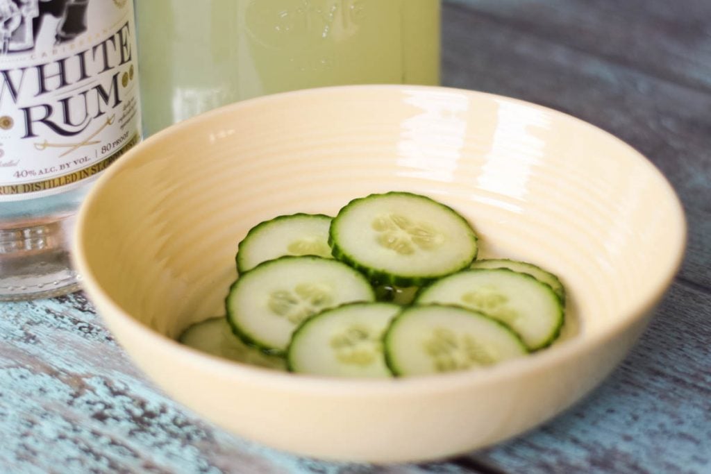 Sliced cucumbers in a shallow bowl with a bottle of white rum in the back of the bowl.