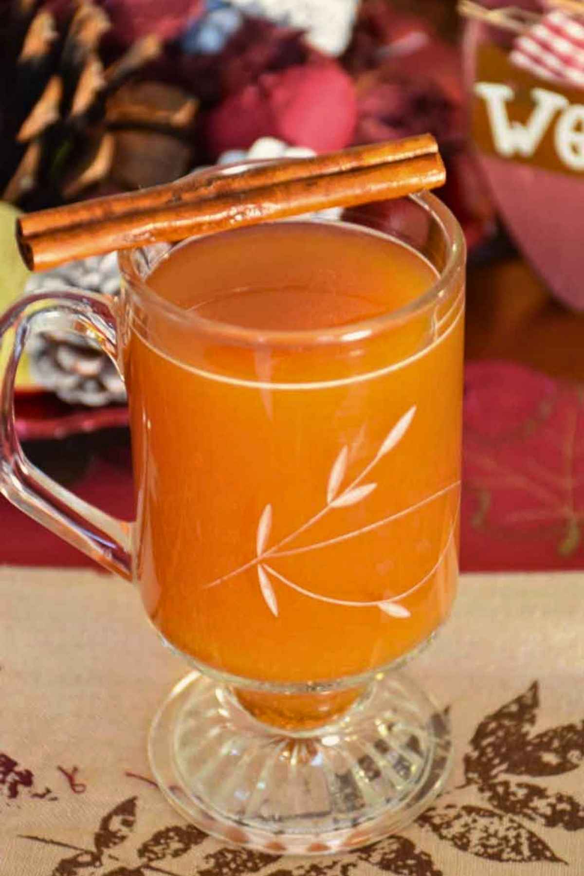 A mug of hot fireball apple cider in a Princess house glas smug with two cinnamon sticks lying across the top of the glass.