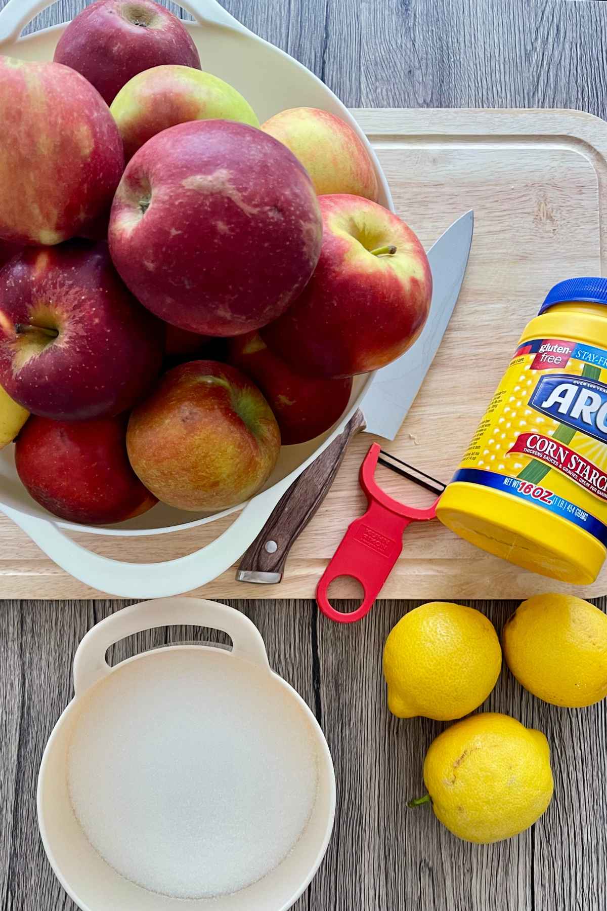 A bowl of red apples, a jar of cornstarch, lemons, and a bowl of sugar set on a wooden board.
