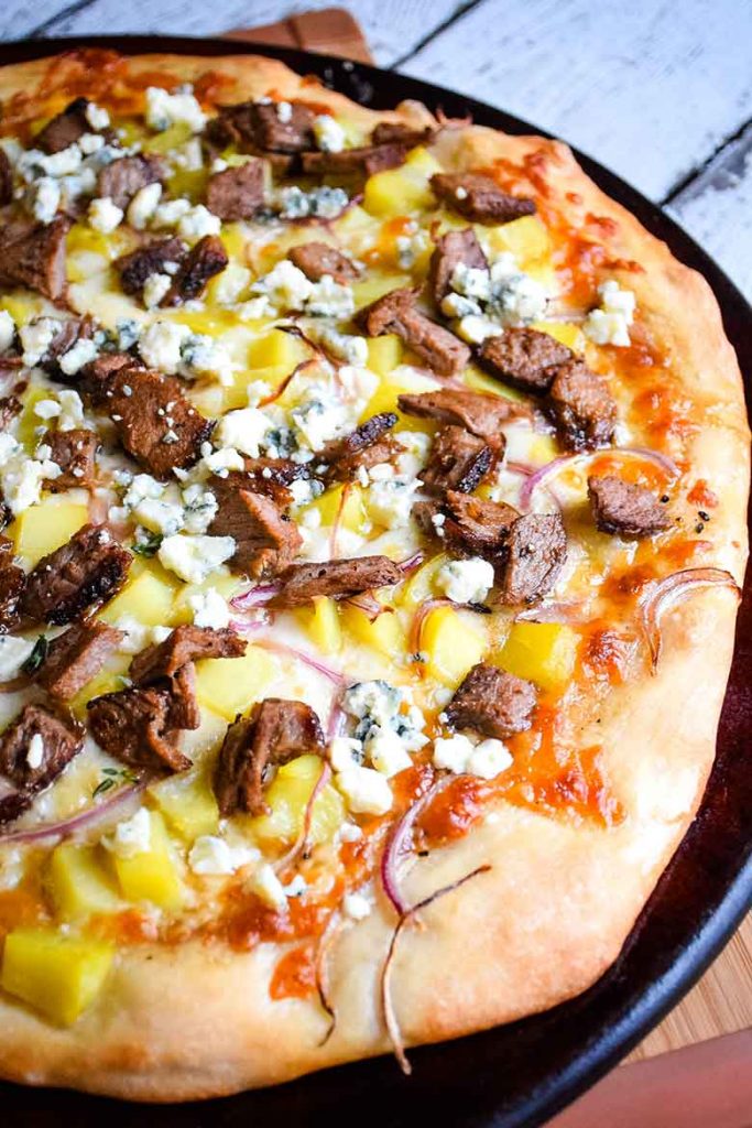 Overhead shot of whole steak and potato pizza on a pizza stone