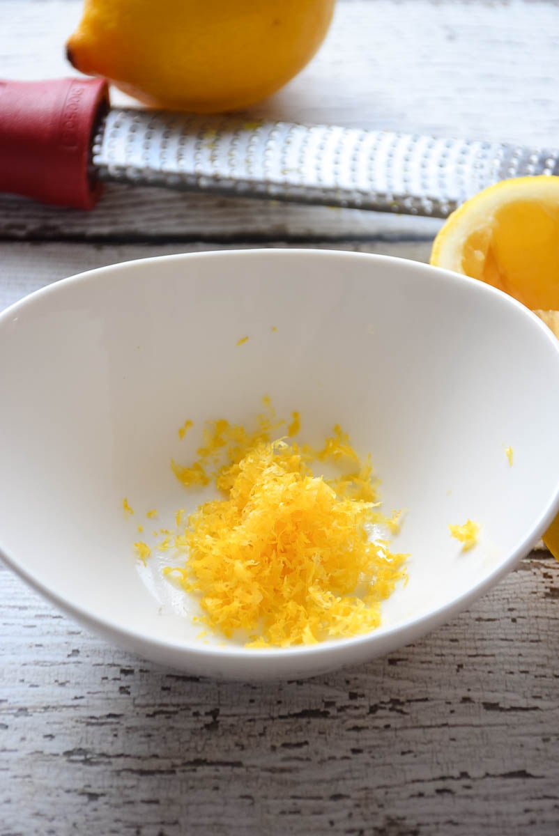 Lemon zest in a white bowl with a microplane zester and lemons in the background