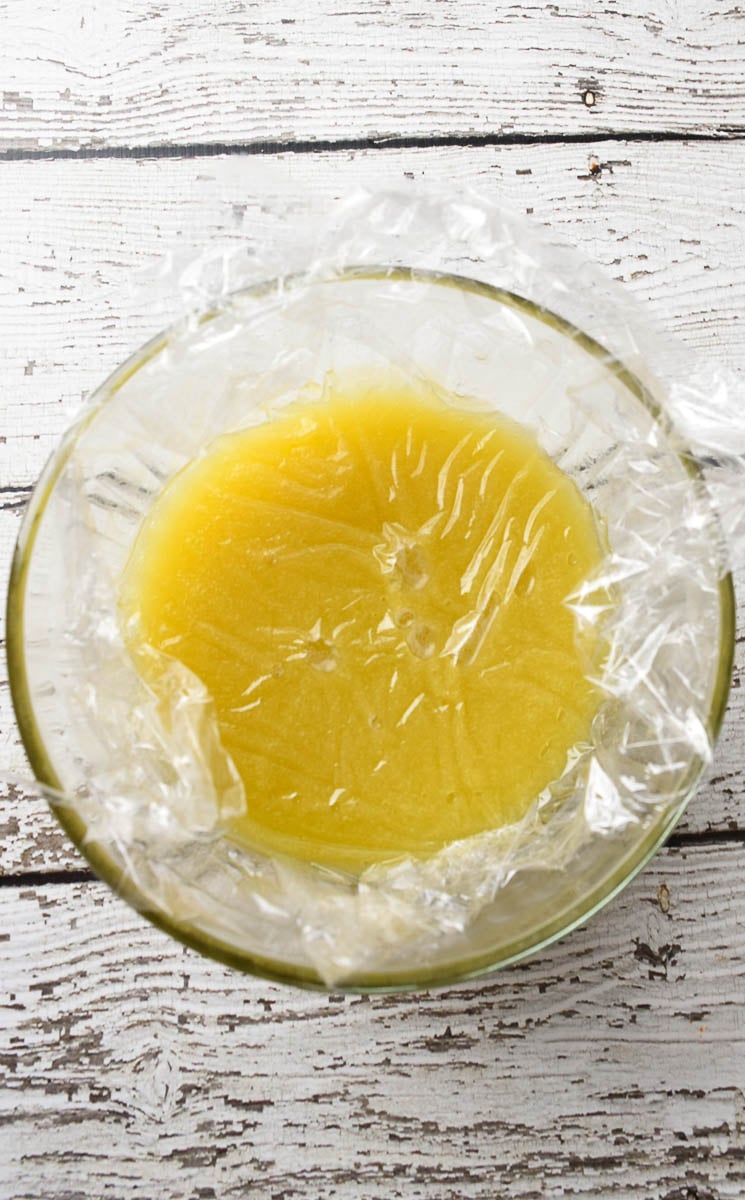 Lemon curd in a glass bowl with plastic wrap pressed into the curd.