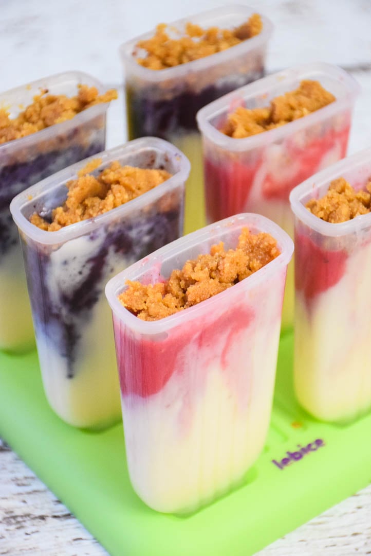 Popsicles in a mold before freezing, topped with crumbled graham cracker crust over visible berry and cheesecake layers.