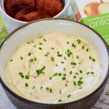 A close-up, overhead view of a creamy white caramelized onion dip in a rustic dark bowl, garnished with fresh chopped chives.