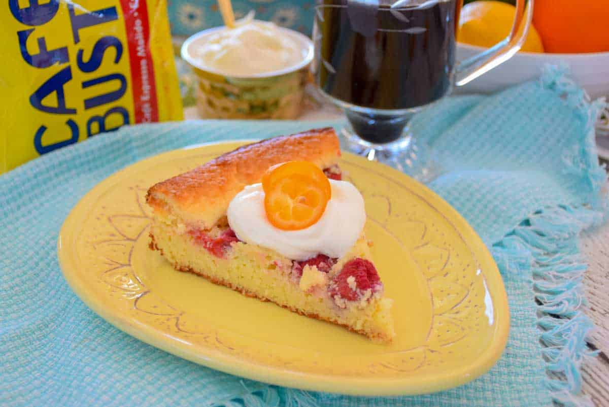 One serving of coffee cake on a yellow egg shaped plate with a blurred glass of coffee blurred in the background to the right and coffee packaging to the left.