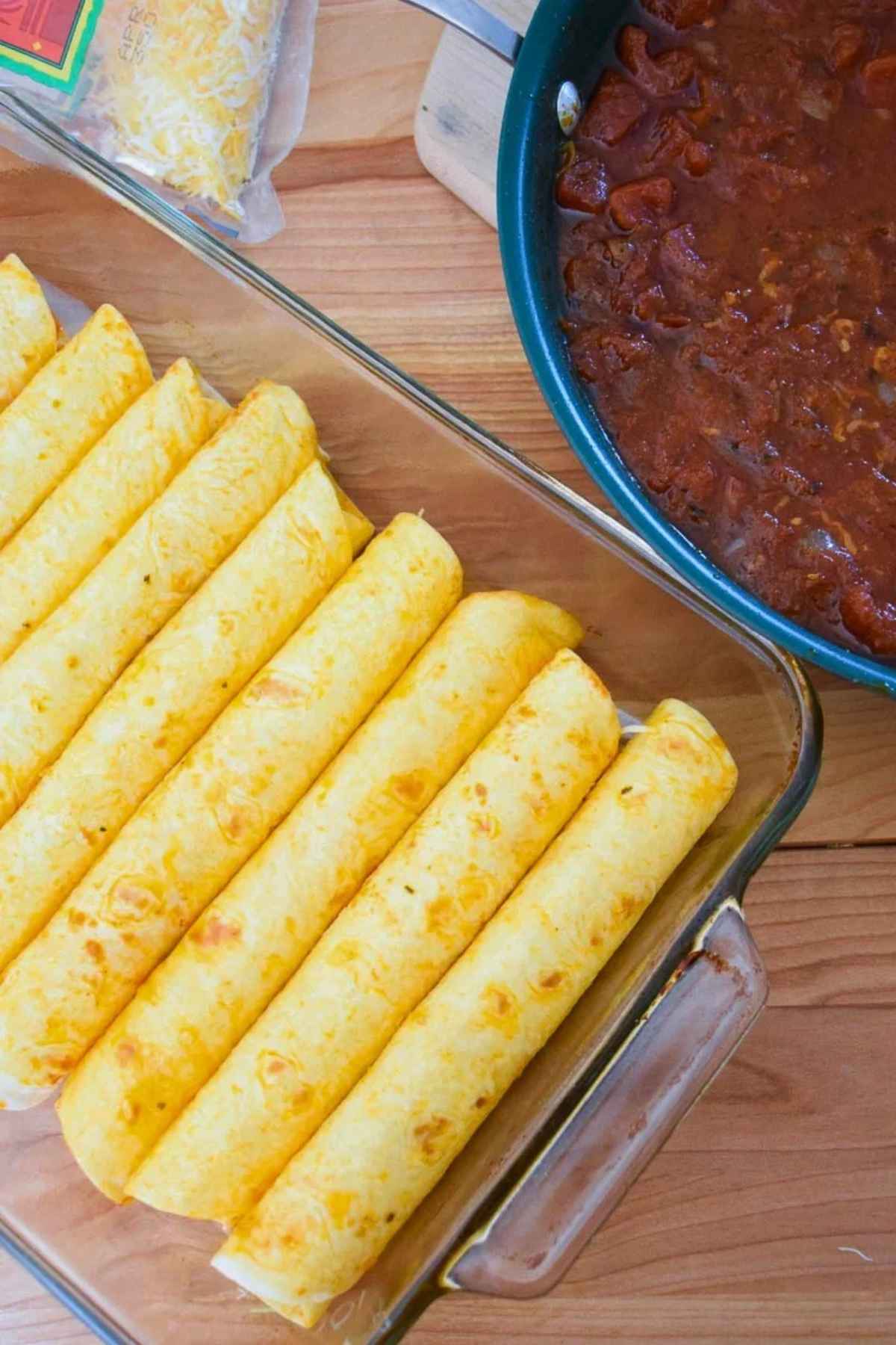 Rolled tortillas lined up in a baking dish next to a skillet and a bag of shredded cheddar.
