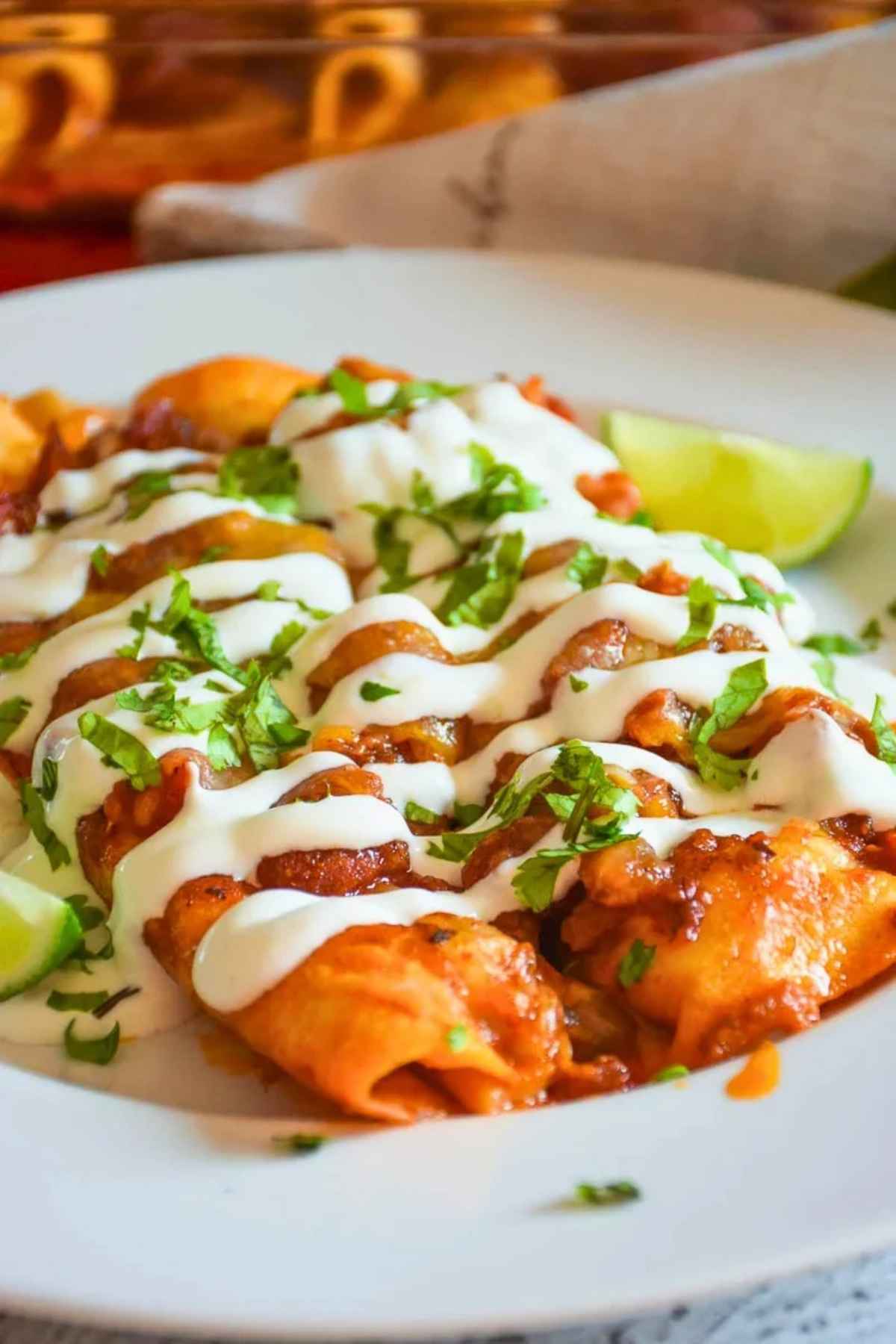 Cheese Enchiladas with Red Sauce drizzled with sour cream on a white plate with lime wedges.