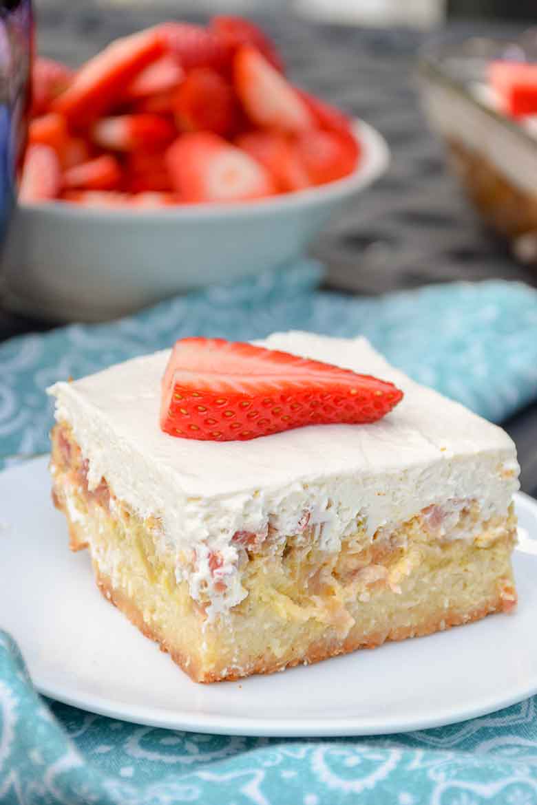 A serving of Keto Rhubarb Dream Bars on a white plate with blue and white napkin and blurred bowl of strawberries in the background