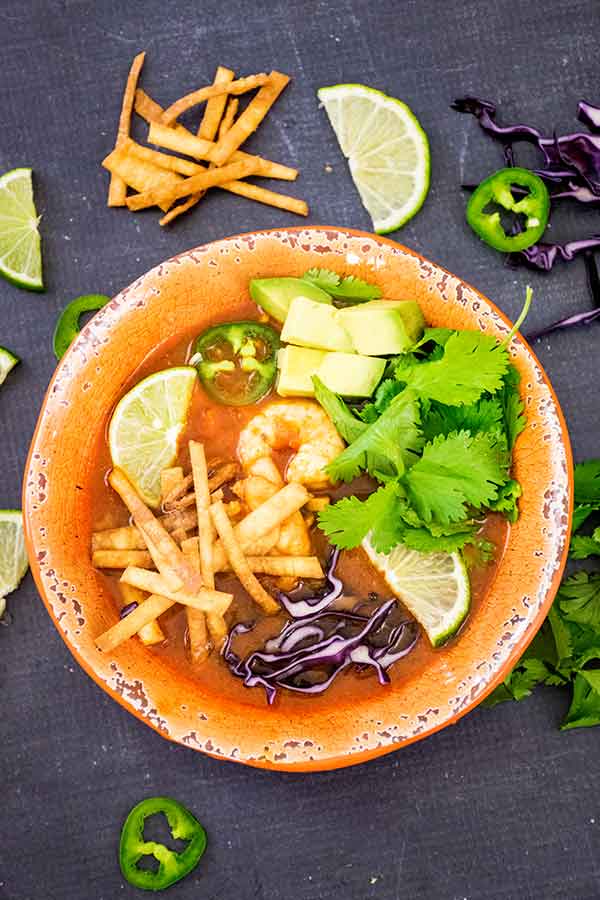 A bowl of shrimp tortilla soup topped with tortilla chips, lime, cilantro, avocado, and jalapenos.