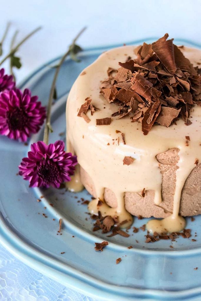 Chocolate Peanut Butter Cheesecake drizzled with sugar free peanut butter sauce and delicately sprinkled with chocolate shavings on a soft blue plate with purple flowers set to the side. 