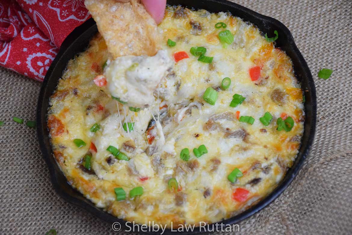 A close-up of a pork rind scooping into cheesy Philly Cheesesteak Dip topped with green onions and diced peppers.