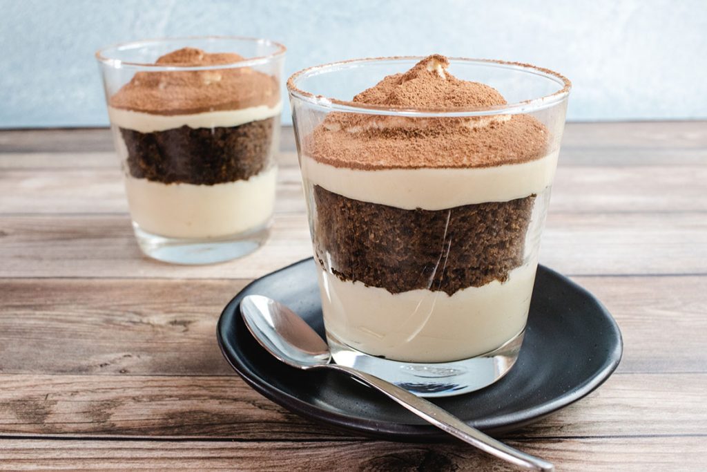 Keto tiramisu in the foreground on a black saucer with a silver spoon on it. There is also another tiramisu in the background. 