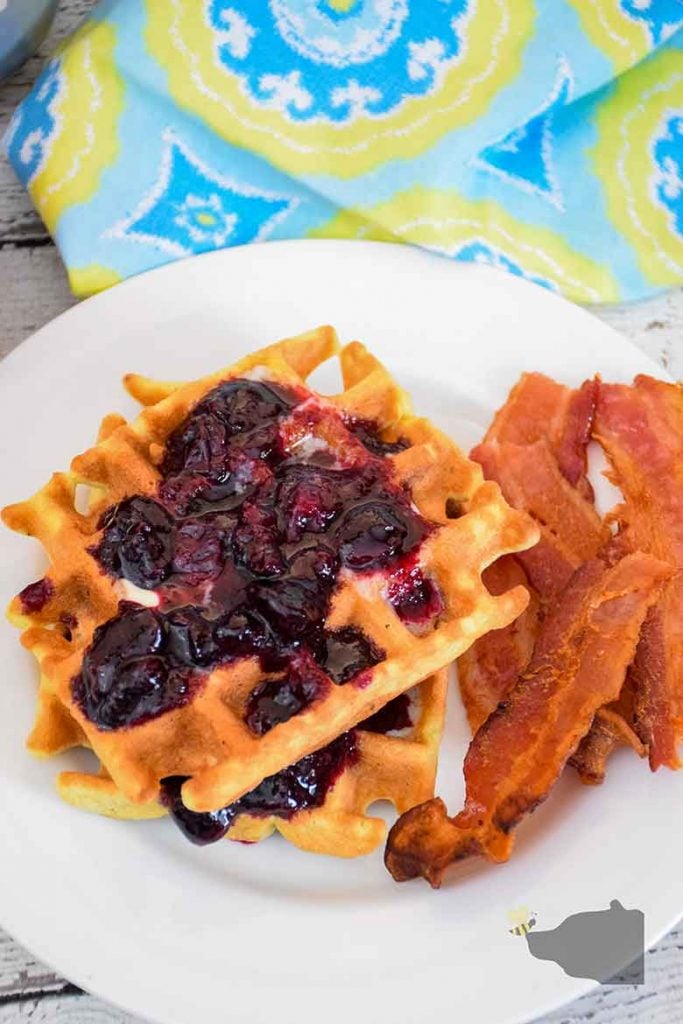 Overhead shot of two blueberry waffles with blueberry sauce and bacon on a white plate