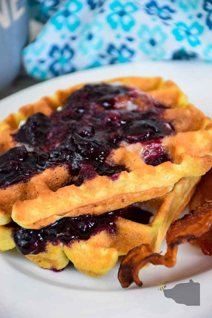 Stack of two blueberry waffles on a white plate with a side of bacon and blue and white napkin