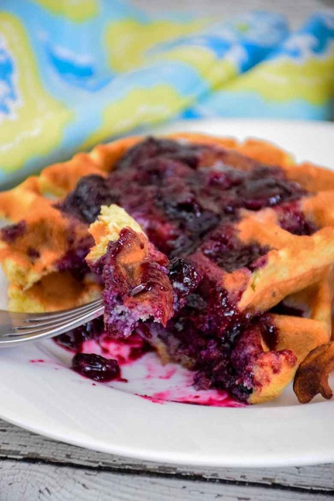 Two cooked keo blueberry waffles on a plate with sugar free blueberry sauce and a section of waffle cut and on a fork