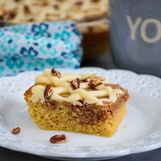 A slice of pecan coffee cake with cinnamon swirl on a white plate with blue and white flowered napkin with baking dish in back and gray coffee mug to the right