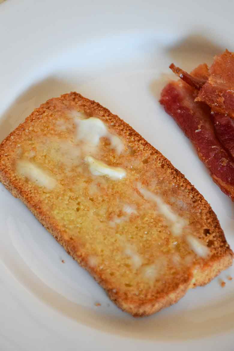 A slice of toasted and buttered grain free bread on a white plate with a side of bacon