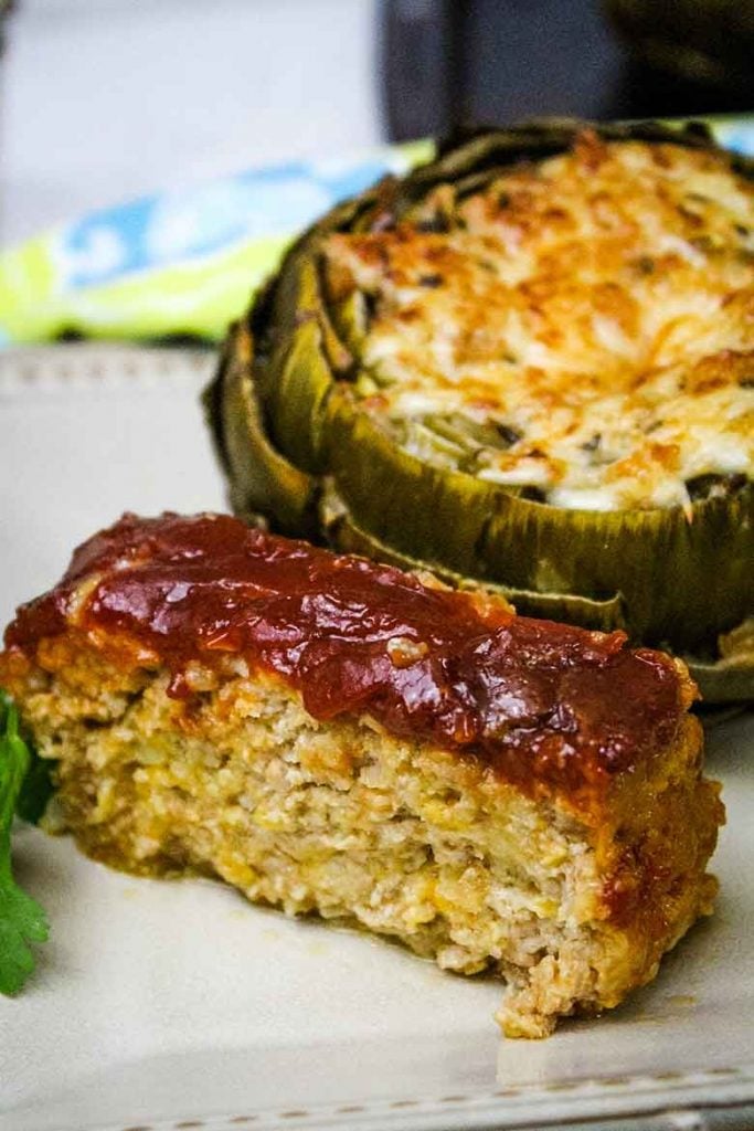 A slice of sweet and sour meatloaf made with ground veal and a stuffed artichoke in the background