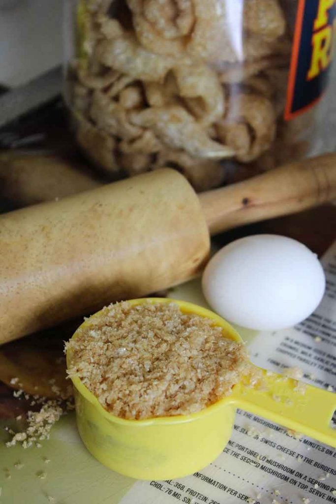 crushed pork rinds in measuring cup with rolling pin, whole egg, and jar of pork rinds in background