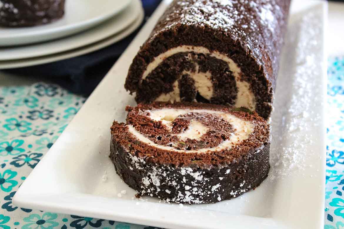 Chocolate Zucchini Cake Roll with a slice on the serving tray and whole roll behind the cake slice