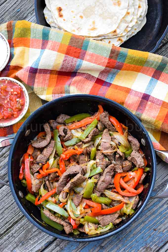 Overhead shot of venison fajitas in a skillet with salsa and flour tortillas in the backgrkound